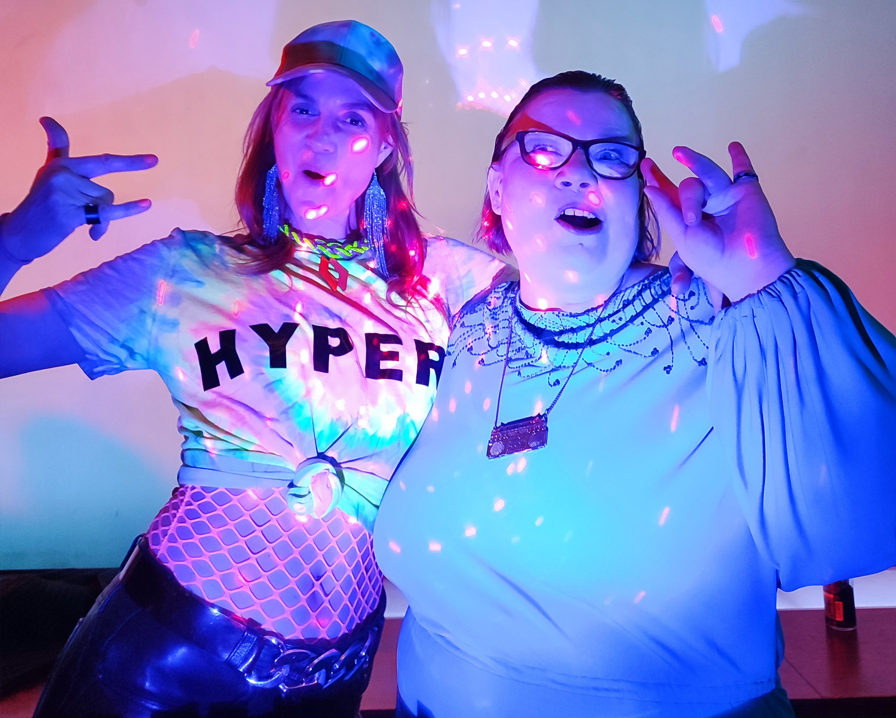 Foto von zwei Personen, die sich in den Armen liegen. Sie werden von bunten Lichtpunkten angeleuchtet. Die linke Person trägt ein T-Shirt mit der Aufschrift "Hyper". Sie strahlen Partylaune aus.