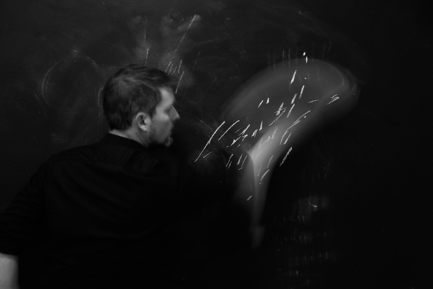 Ein Mann im schwarzen Hemd und kurzen dunklen Haaren mit dem Rücken zur Kamera. Er schreibt oder malt mit Kreide an einer Tafel. Sein Arm bewegt sich sehr schnell und ist unscharf.