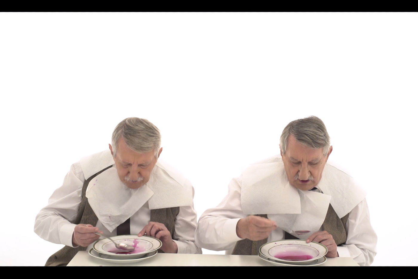 Zwei Männer mit grauen Haaren und Schnauzbärten an einem Tisch. Beide tragen weiße Hemden und Papierservietten um den Hals. Sie sehen sich sehr ähnlich, wie Zwillinge. Sie essen pinke Suppe aus Tellern vor sich.