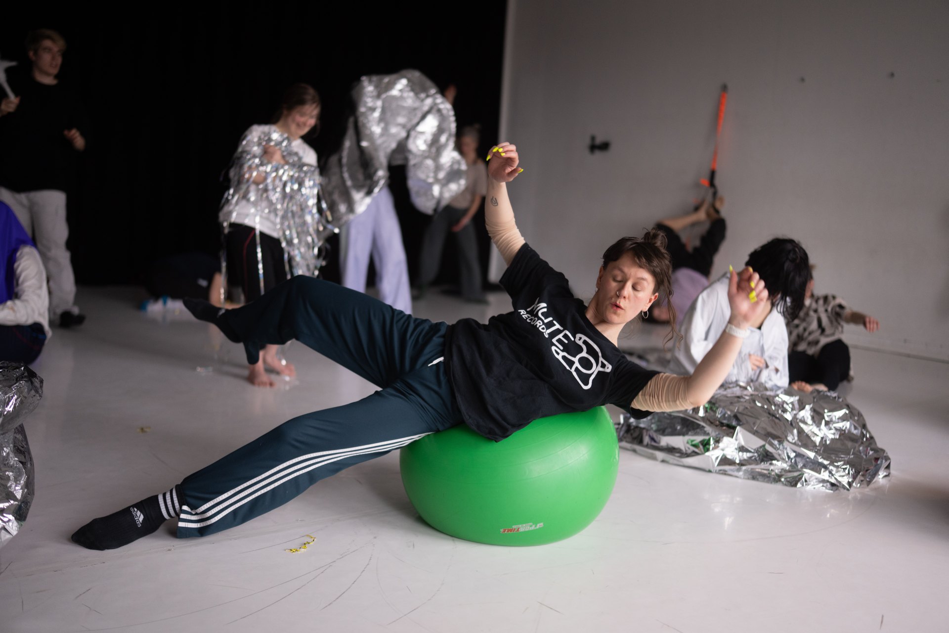 Eine Person in dunkler Sportkleidung balanciert seitlich liegend auf einem leuchtend grünen Gymnastikball. Sie hat die Arme und ein Bein ausgestreckt, um die Balance zu halten. Im Hintergrund sind weitere Personen im Raum verteilt, die teilweise mit große
