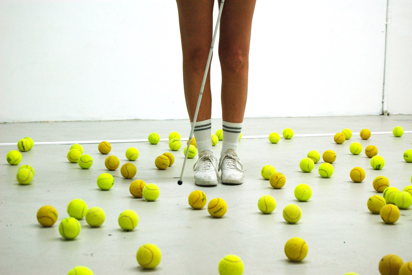 Die Beine einer Person. Sie trägt weiße Schuhe und Tennissocken, und einen weißen Stock. Um sie herum liegen viele gelbe Tennisbälle auf dem Boden.