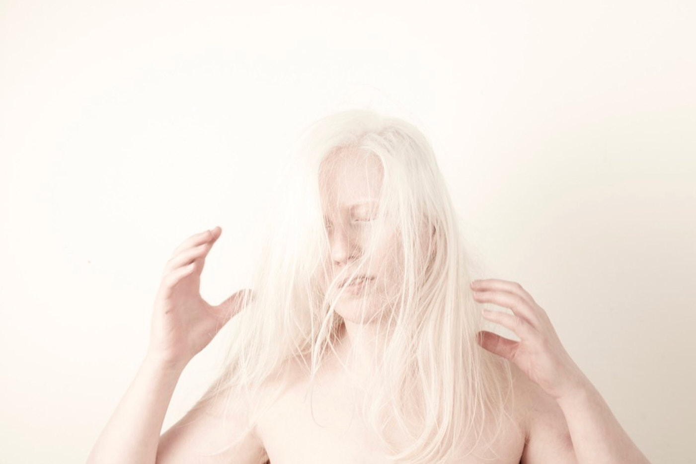 Eine Person mit langen blonden Haaren in hellem Licht vor einem hellen Hintergrund. Sie hält ihre Hände angewinkelt in die Luft, wie um etwas zu greifen. Die Haare verdecken teilweise ihr Gesicht.