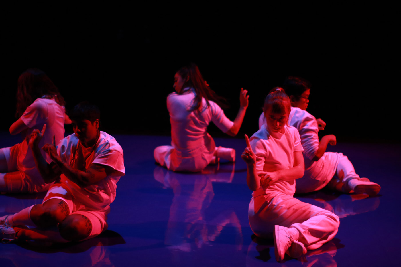 Fünf Personen in weißer Kleidung sitzen im Kreis auf einem blauen Tanzboden. Sie schauen nach außen und haben ihre Arme zu Gesten angewinkelt. Rotes Licht scheint auf sie. 