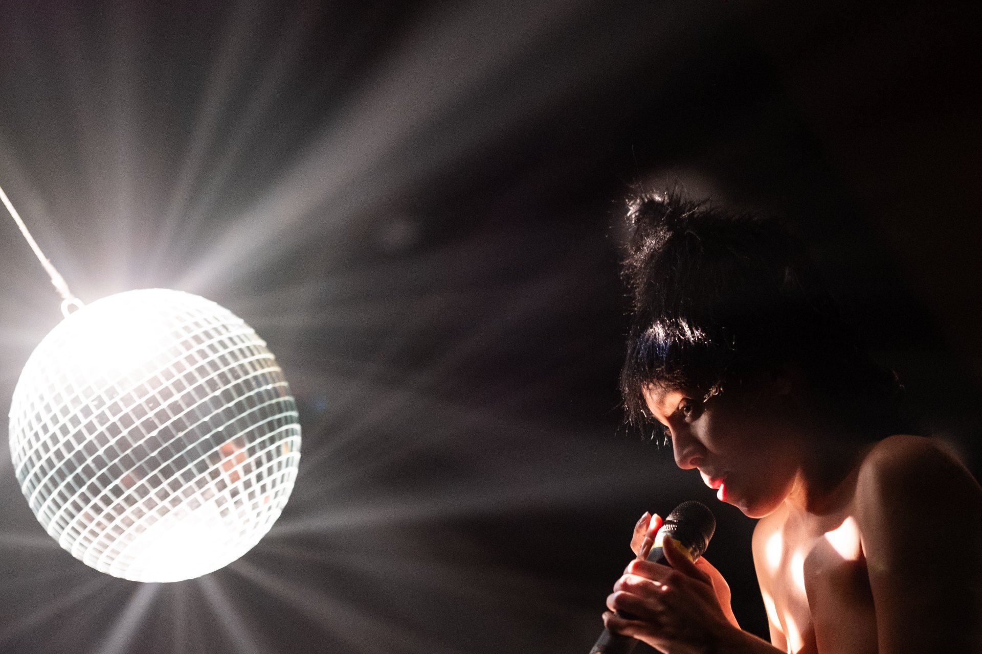 Eine Person ist im Profil zu sehen, wie sie ein Mikrofon nah an den Mund hält und konzentriert nach unten blickt. Links im dunklen Raum hängt eine hell erleuchtete Discokugel, die intensive weiße Lichtstrahlen in die Umgebung wirft.